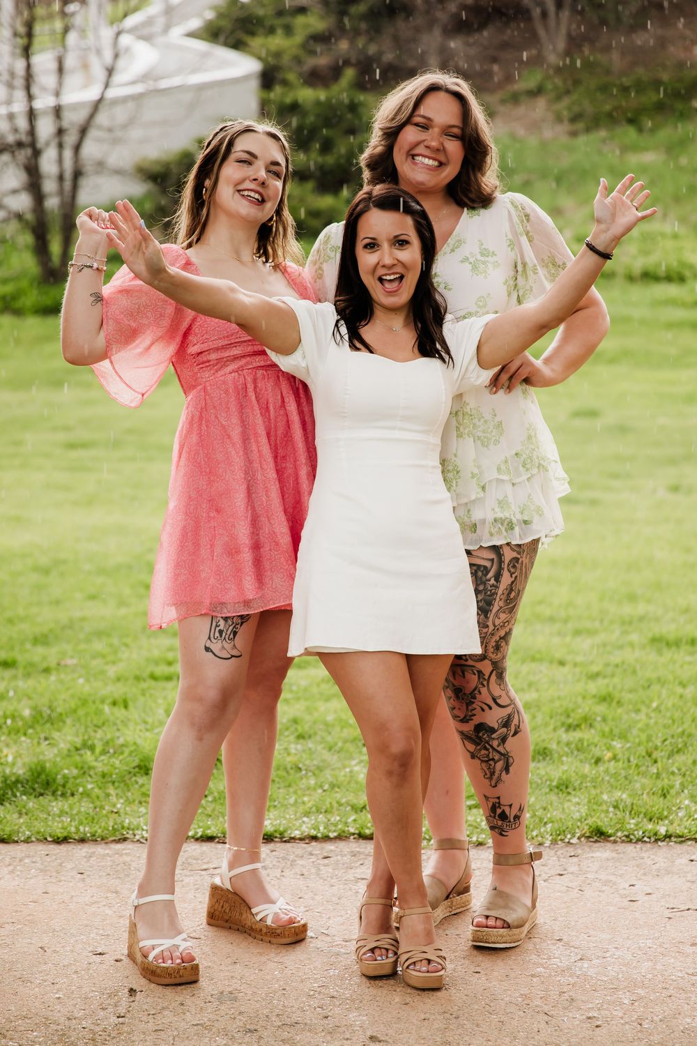 Friends in pink and white dresses pose together playfully on a grassy path.