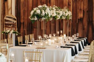Elegant wedding reception table with tall floral centerpiece and gold chiavari chairs in rustic barn venue.