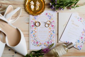 Wedding invitation with floral design surrounded by rings, shoes, and decorative elements on wooden surface.