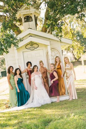 Wedding party in colorful dresses poses together by white chapel gazebo.
