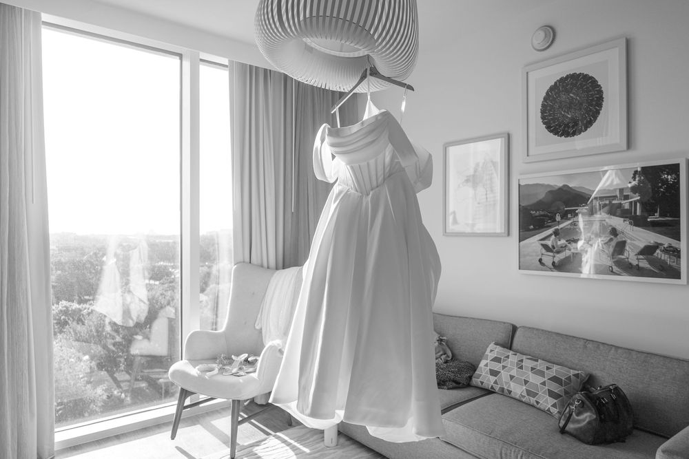 Black and white photo of a white wedding dress hanging near a large window in a modern hotel room.
