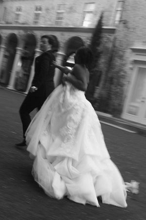 Black and white photo of a bride in a voluminous white gown and groom in a suit walking briskly outdoors.