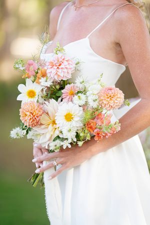 A delicate bridal bouquet featuring peachy-pink dahlias, white daisies and soft green foliage in natural light.