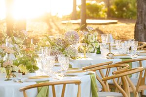 Elegant outdoor wedding reception table with white linens, wooden chairs and glowing golden sunlight through trees.