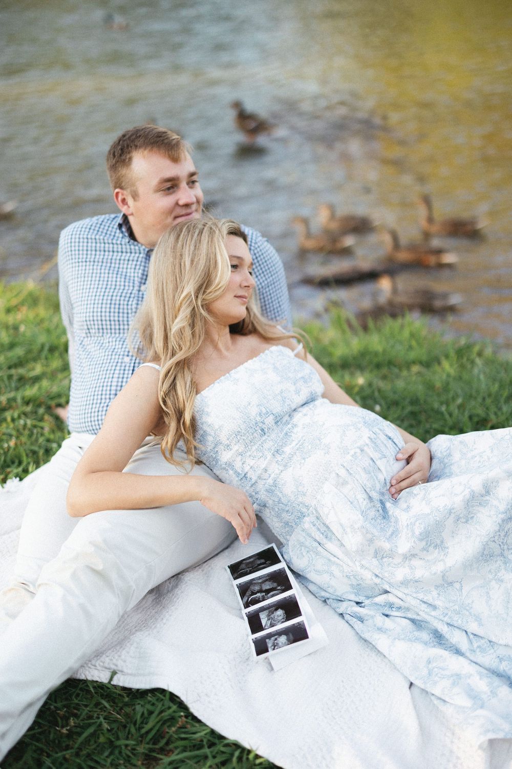 Pregnant couple sitting by a pond holding ultrasound photos, woman in blue floral dress, man in plaid shirt, ducks in background.