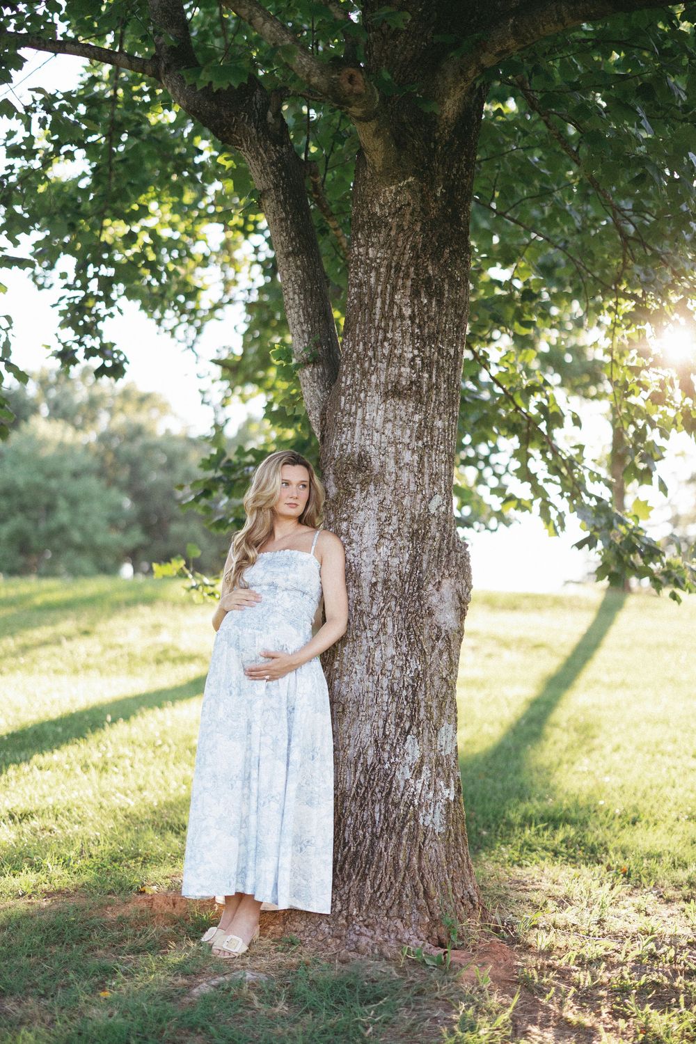 Pregnant woman in floral dress leaning against large tree in sunny green park, cradling baby bump.