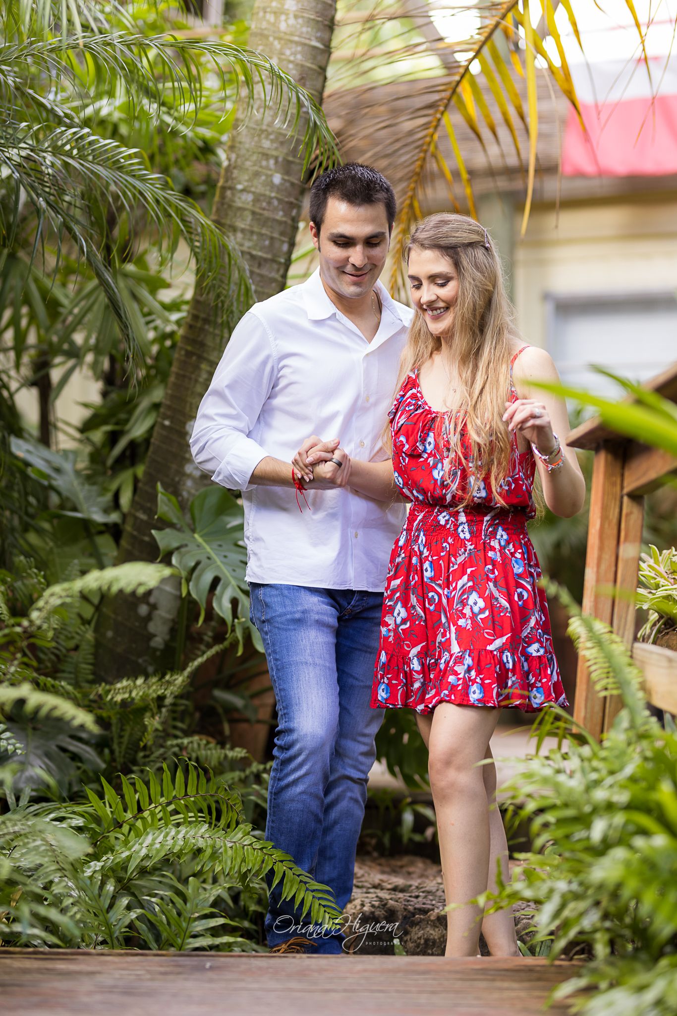 Couple walking hand in hand on wooden path surrounded by lush tropical greenery, man in white shirt and woman in red dress.