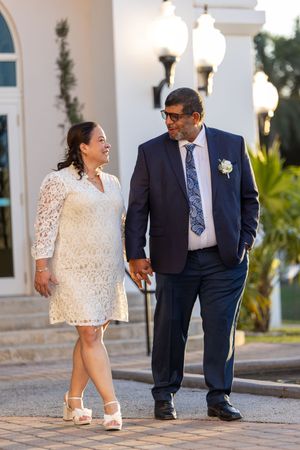 Couple in wedding attire smiling at each other outside elegant venue, woman in white lace dress, man in navy suit.