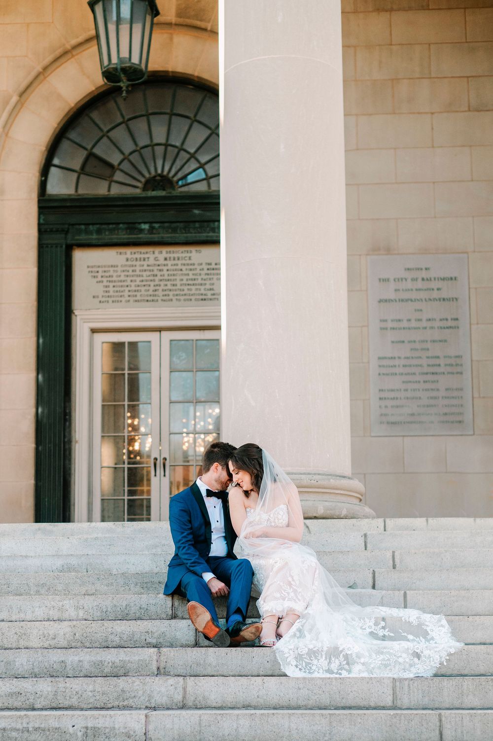 A romantic couple in wedding attire sits on grand stone steps of a historic building with elegant columns.