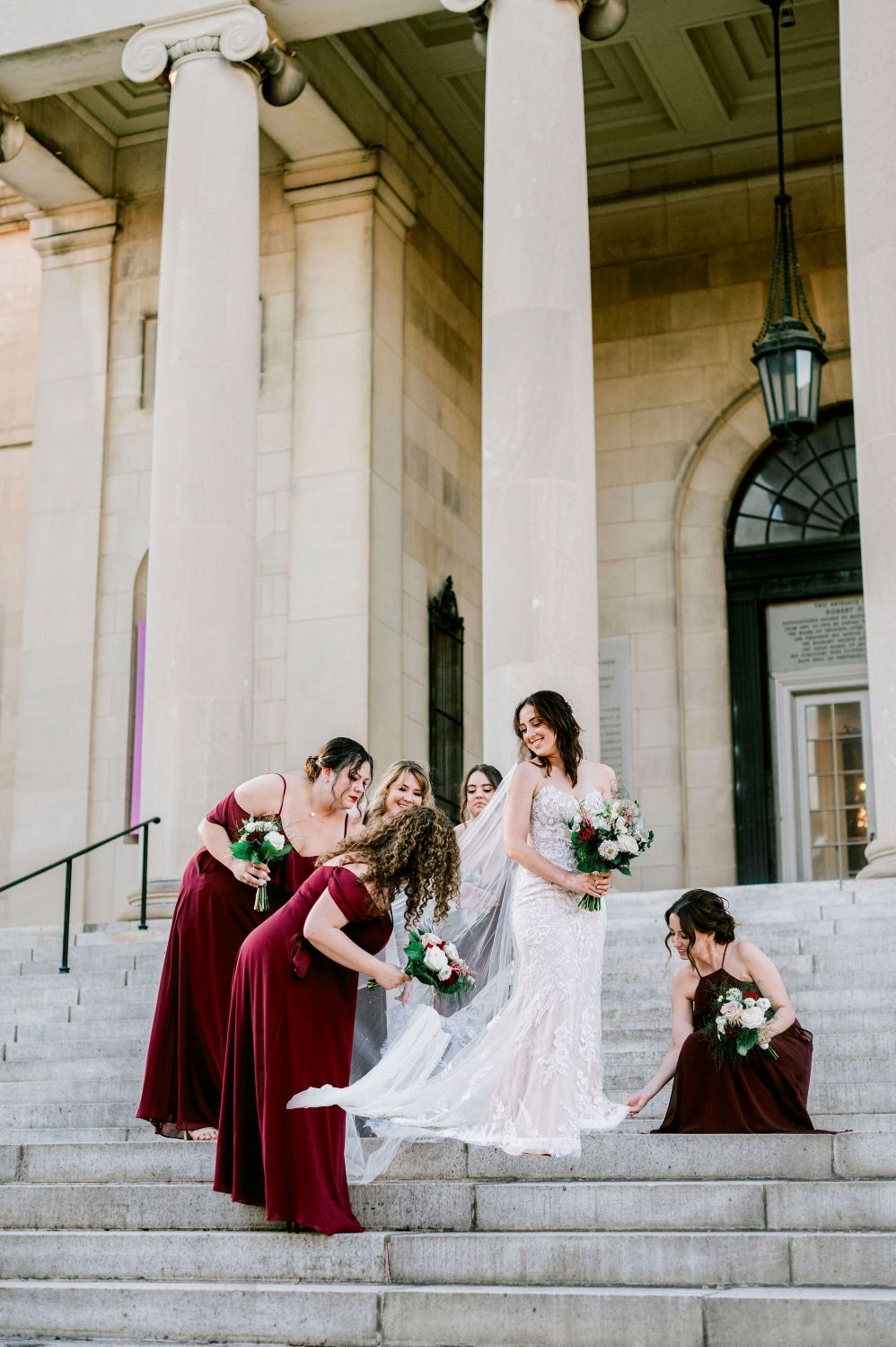 Wedding party in burgundy dresses poses playfully on white stone steps with columns in the background.