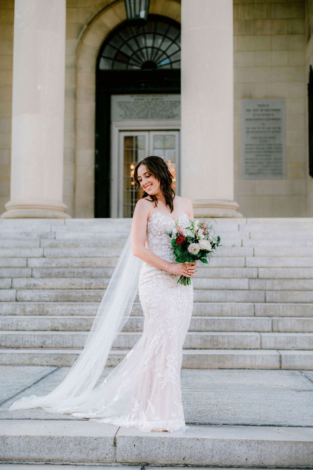A bride in a lace wedding gown poses elegantly on grand white marble steps of a classical building.