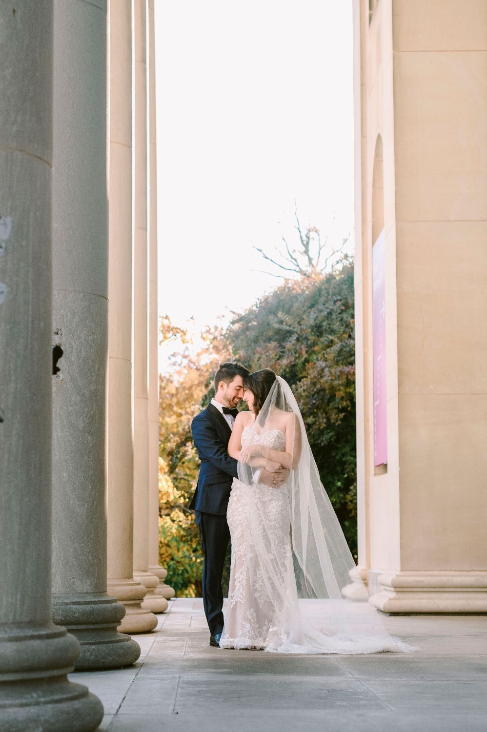 Romantic wedding couple shares intimate moments under elegant stone columns during sunset photo session.