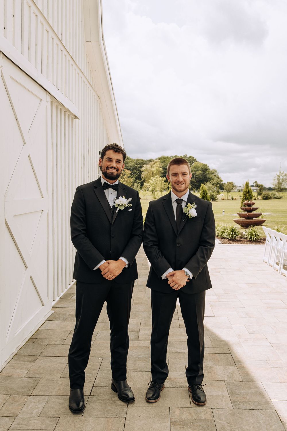 Two groomsmen in black tuxedos with boutonnieres standing outside a white barn wedding venue on a cloudy day.