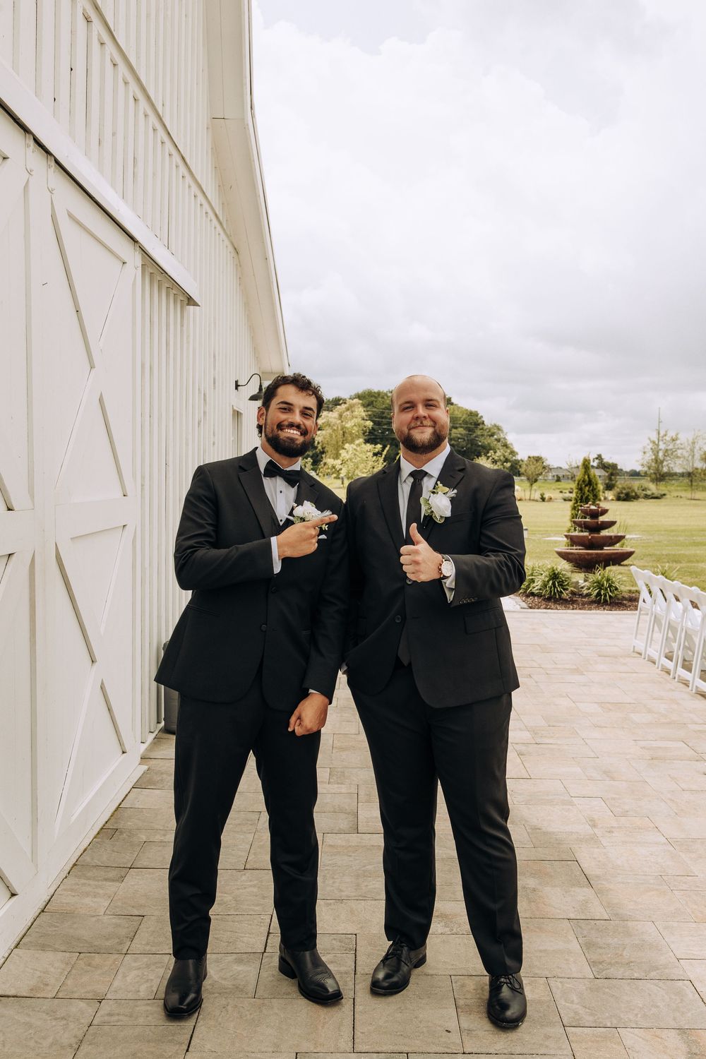 Two groomsmen in black tuxedos with white boutonnieres pose with thumbs up outside a white barn wedding venue.