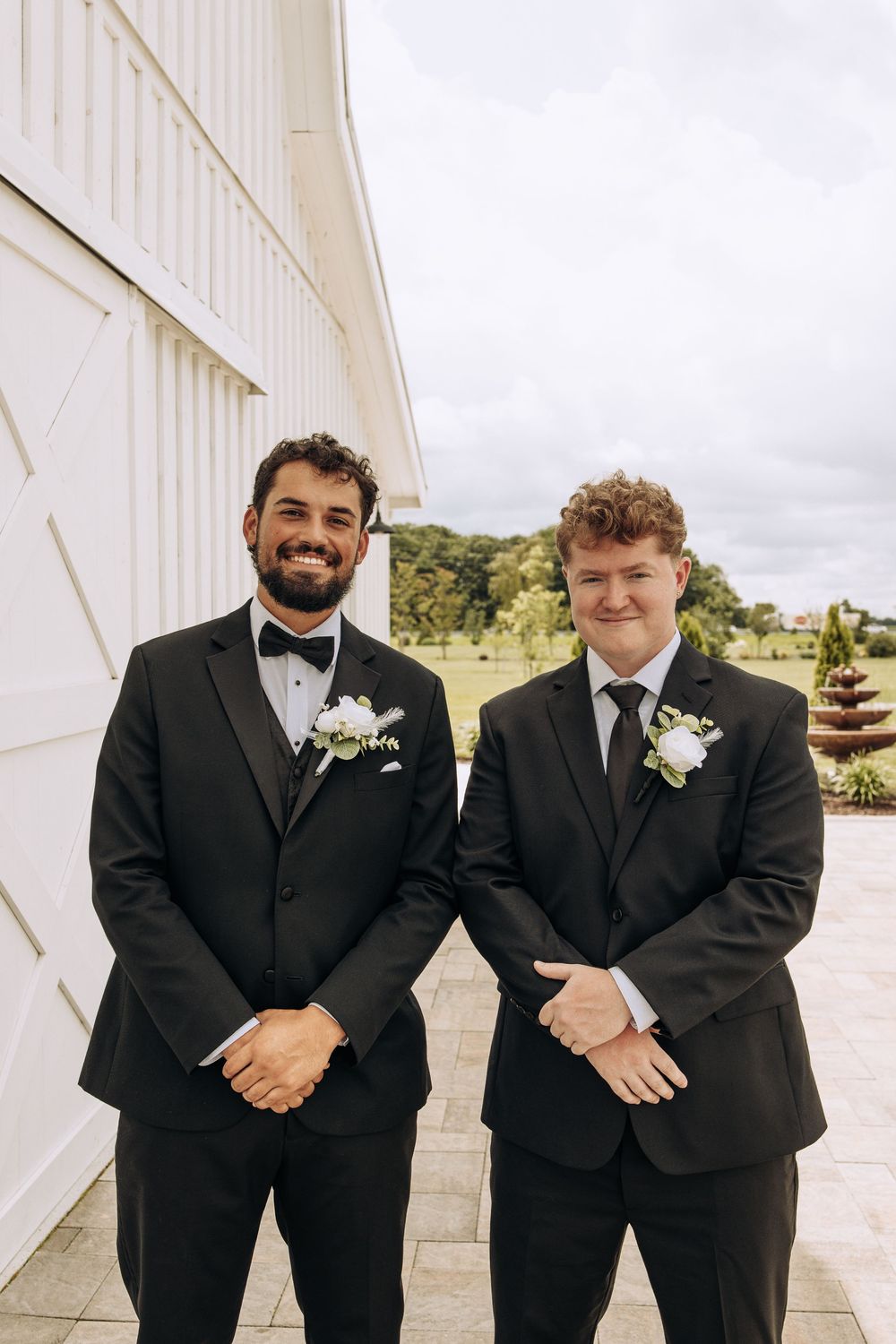 Two groomsmen in black suits with white boutonnieres smile outside a white barn wedding venue on a cloudy day.