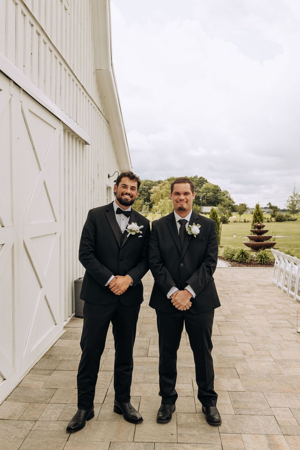 Two groomsmen in black tuxedos with white boutonnieres stand smiling outside a white barn wedding venue.