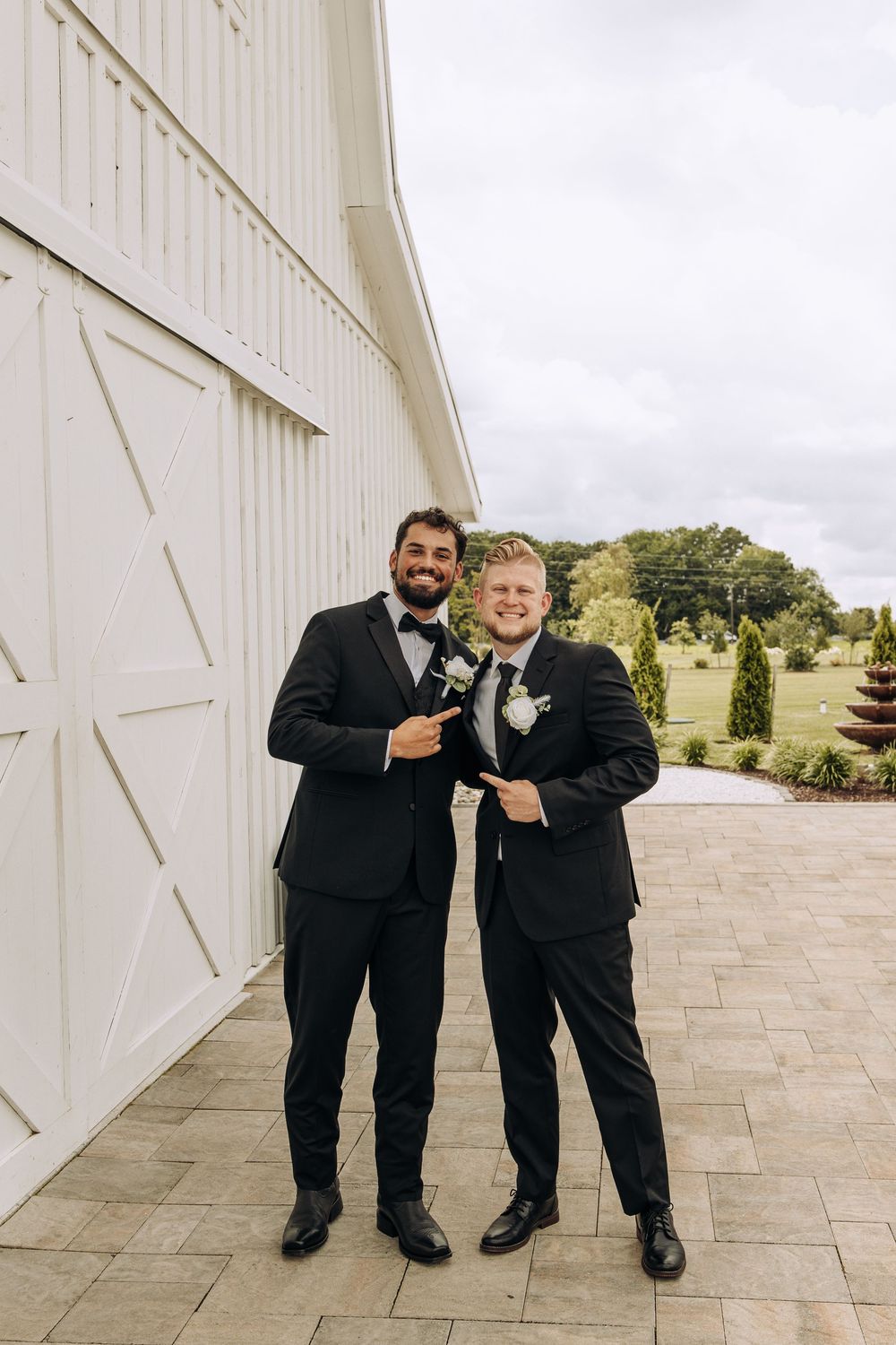 Two groomsmen in black tuxedos with white boutonnieres point at each other smiling outside a white barn venue.