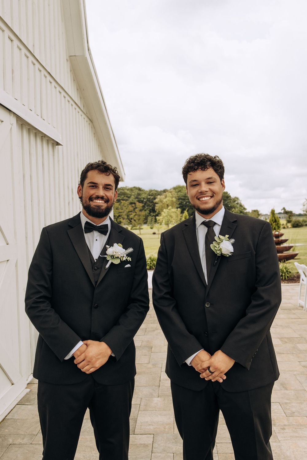 Two groomsmen in black suits with white boutonnieres smile together outside a white barn wedding venue.