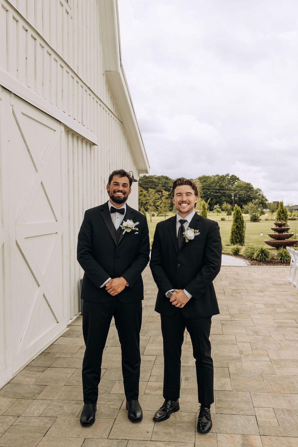 Two groomsmen in black tuxedos with white boutonnieres pose smiling outside a white barn wedding venue.