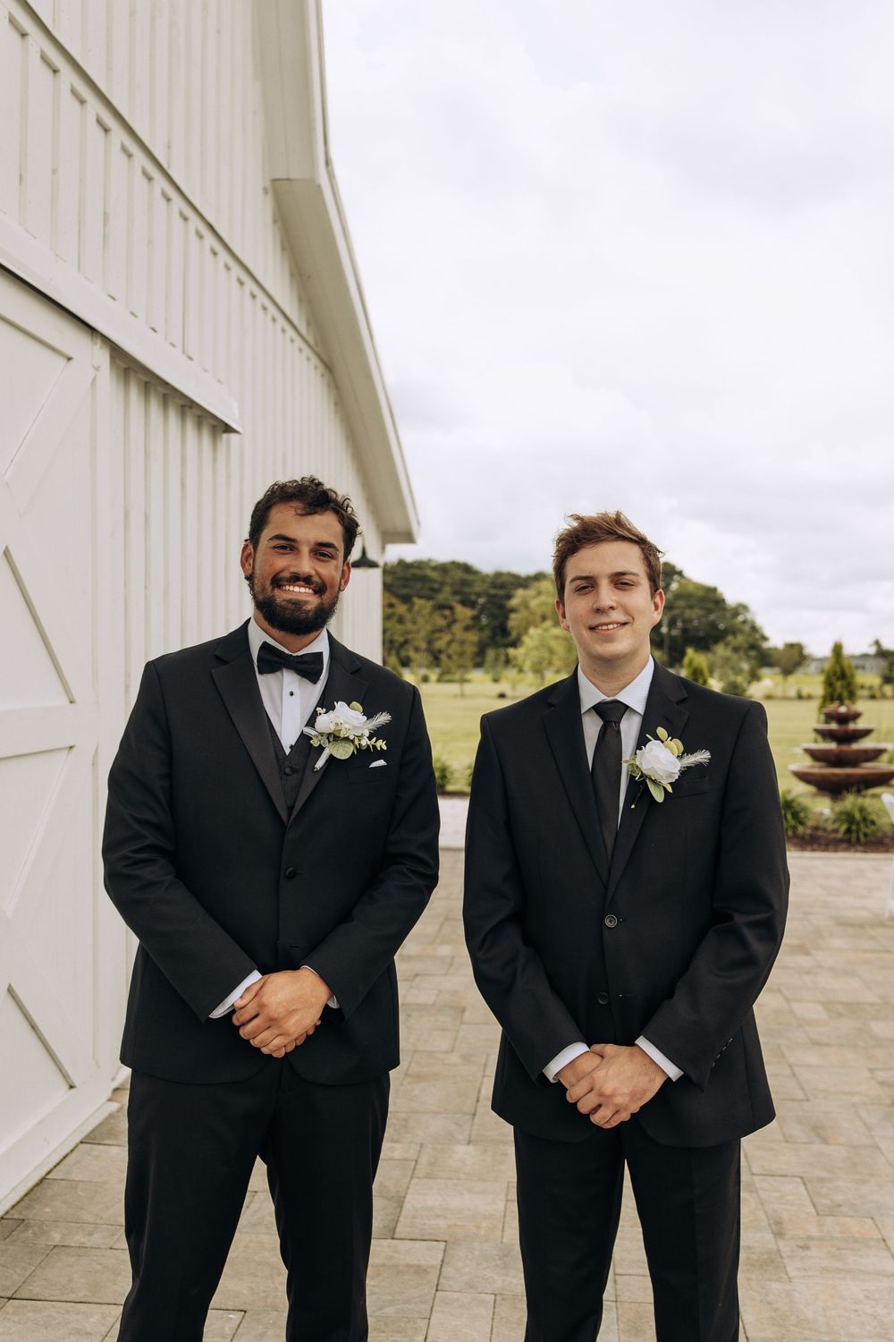 Two groomsmen in black suits with white boutonnieres stand smiling outside a white barn wedding venue.