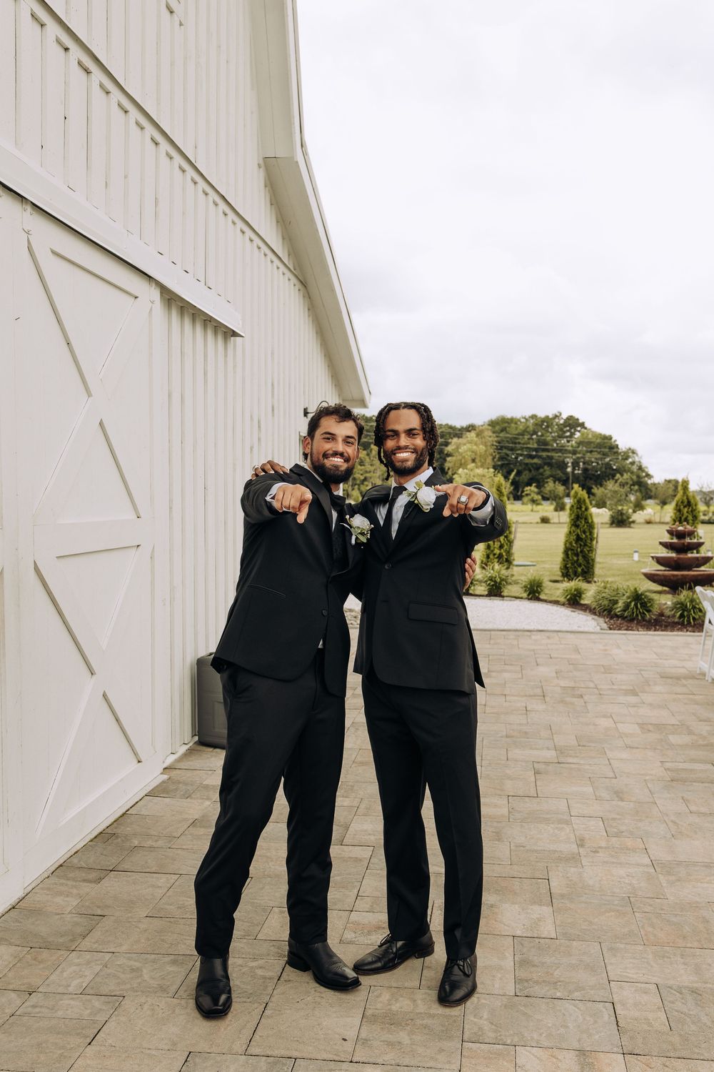 Two groomsmen in black tuxedos embrace and point at camera outside a white barn wedding venue.