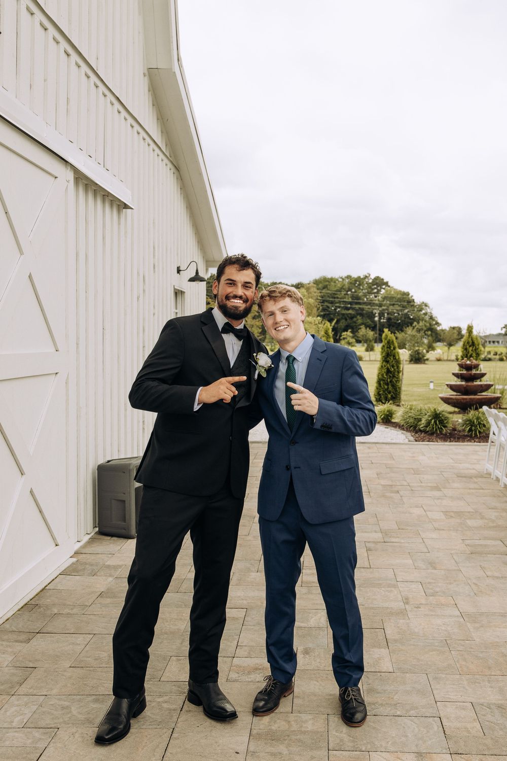 Two groomsmen in black and navy suits with boutonnieres point at each other outside a white barn venue.