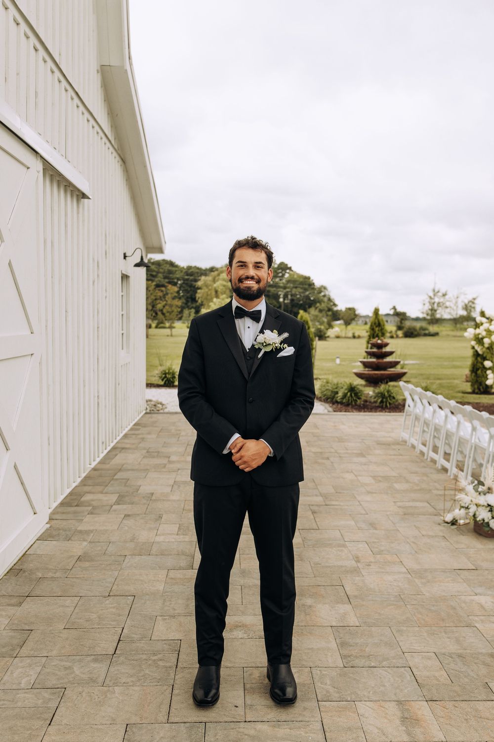 Smiling groom in black tuxedo with bow tie and boutonniere stands on stone patio outside white barn venue.