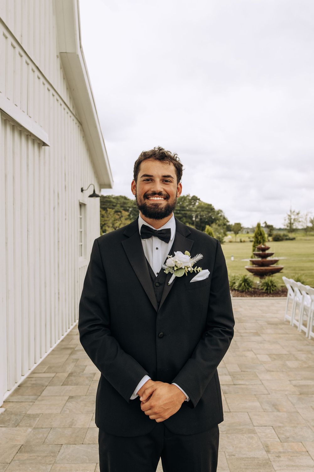 Handsome groom in black tuxedo smiles warmly wearing white floral boutonniere outside elegant barn wedding venue.