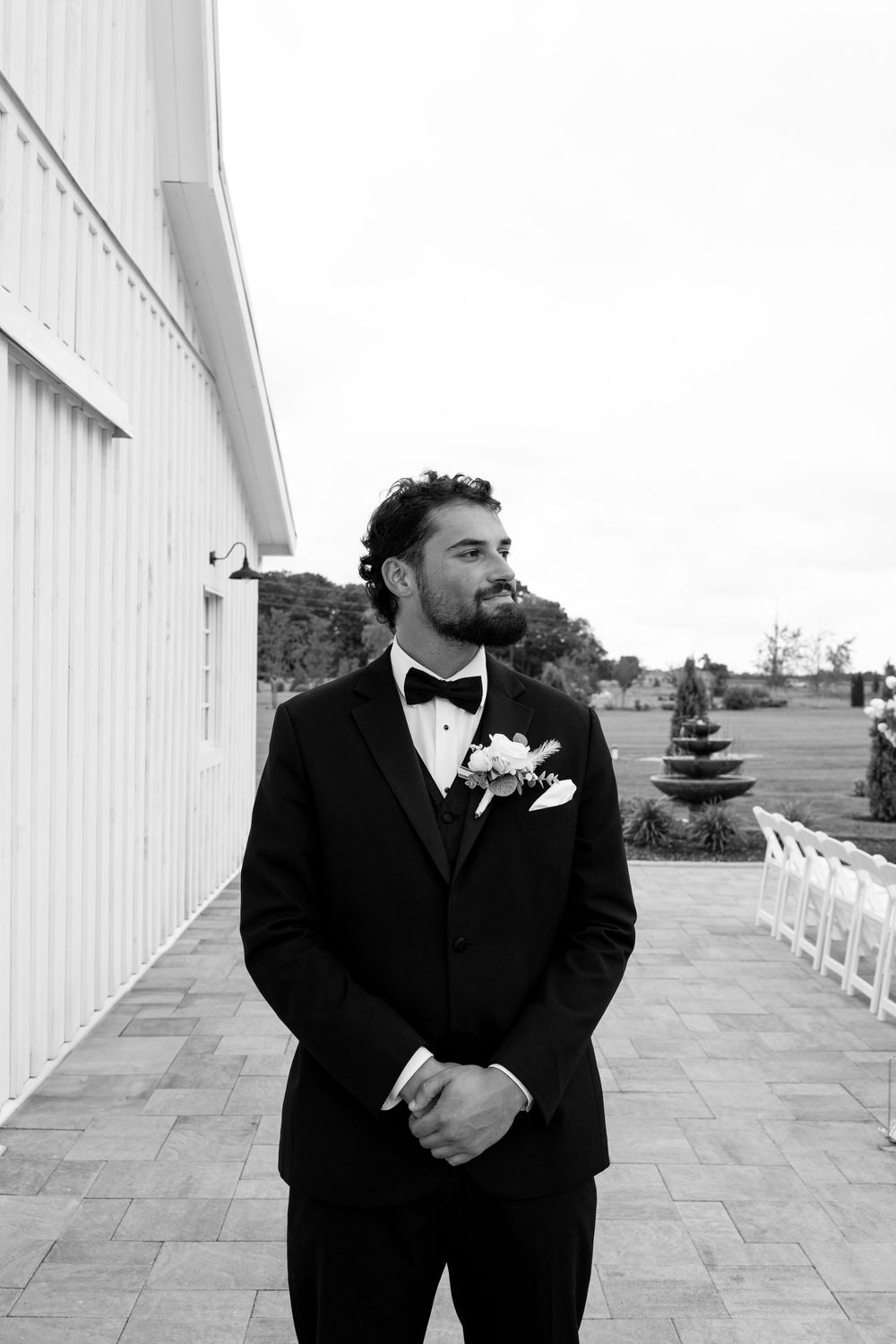 Black and white portrait of groom in tuxedo with boutonniere gazing thoughtfully beside white barn wedding venue.
