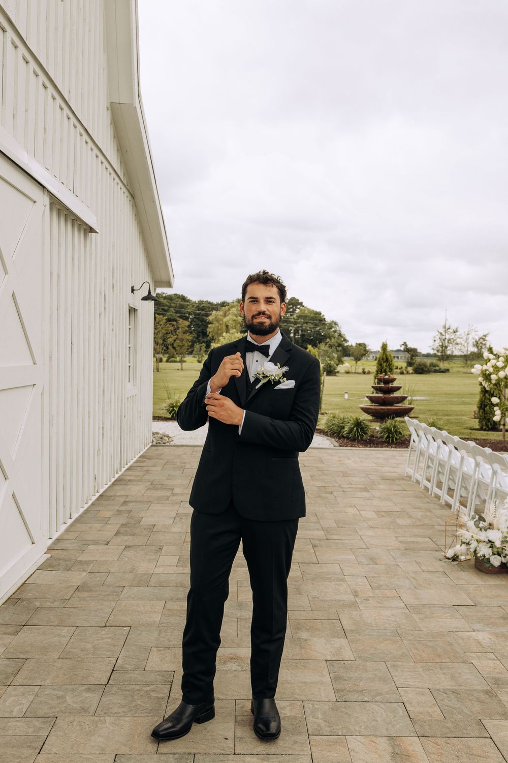 Dapper groom in black tuxedo adjusting cufflinks on stone patio outside white barn wedding venue with green grounds.