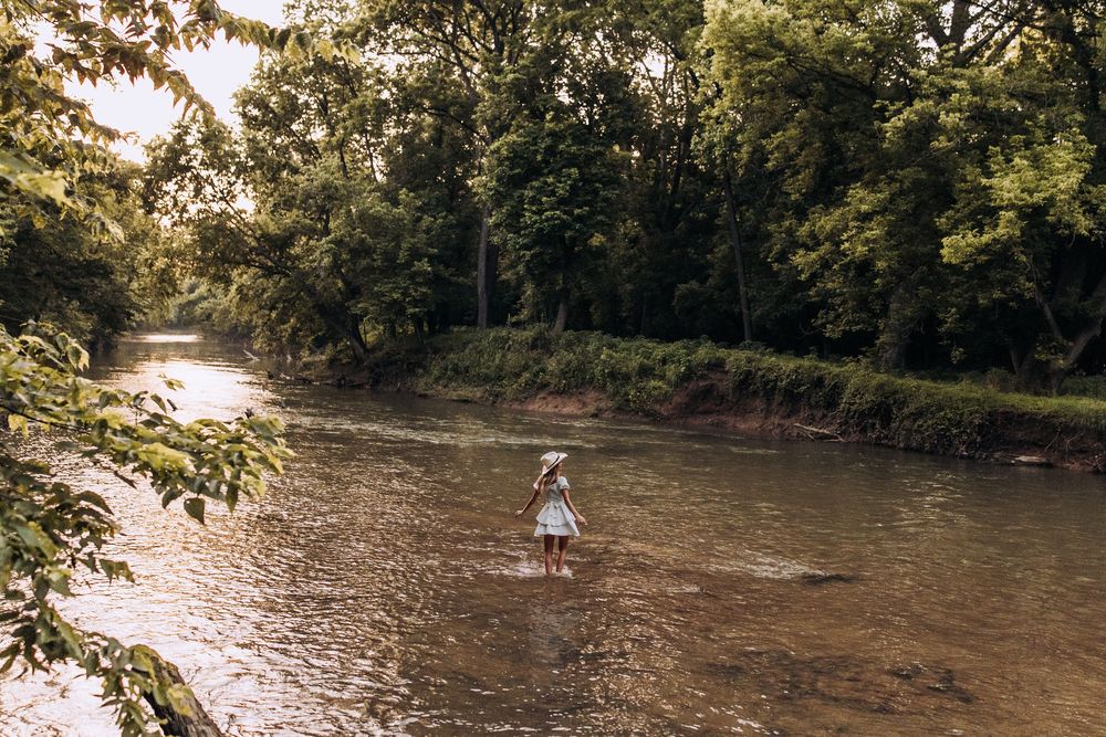 Woman in white dress and hat wades through a shallow tree-lined river.