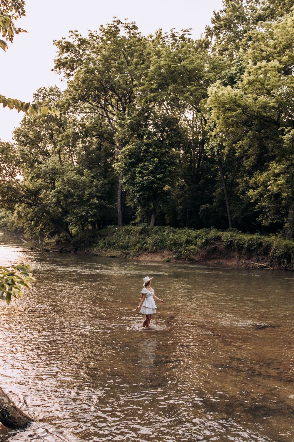 Woman in white dress and hat stands in a shallow river surrounded by lush green trees.