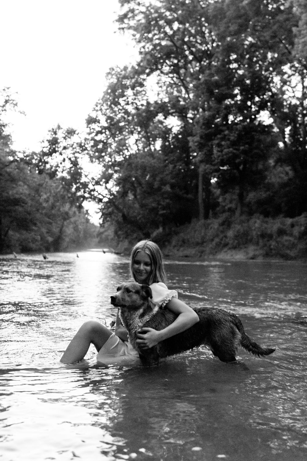 Black and white photo of a smiling woman hugging her dog while sitting in a river.