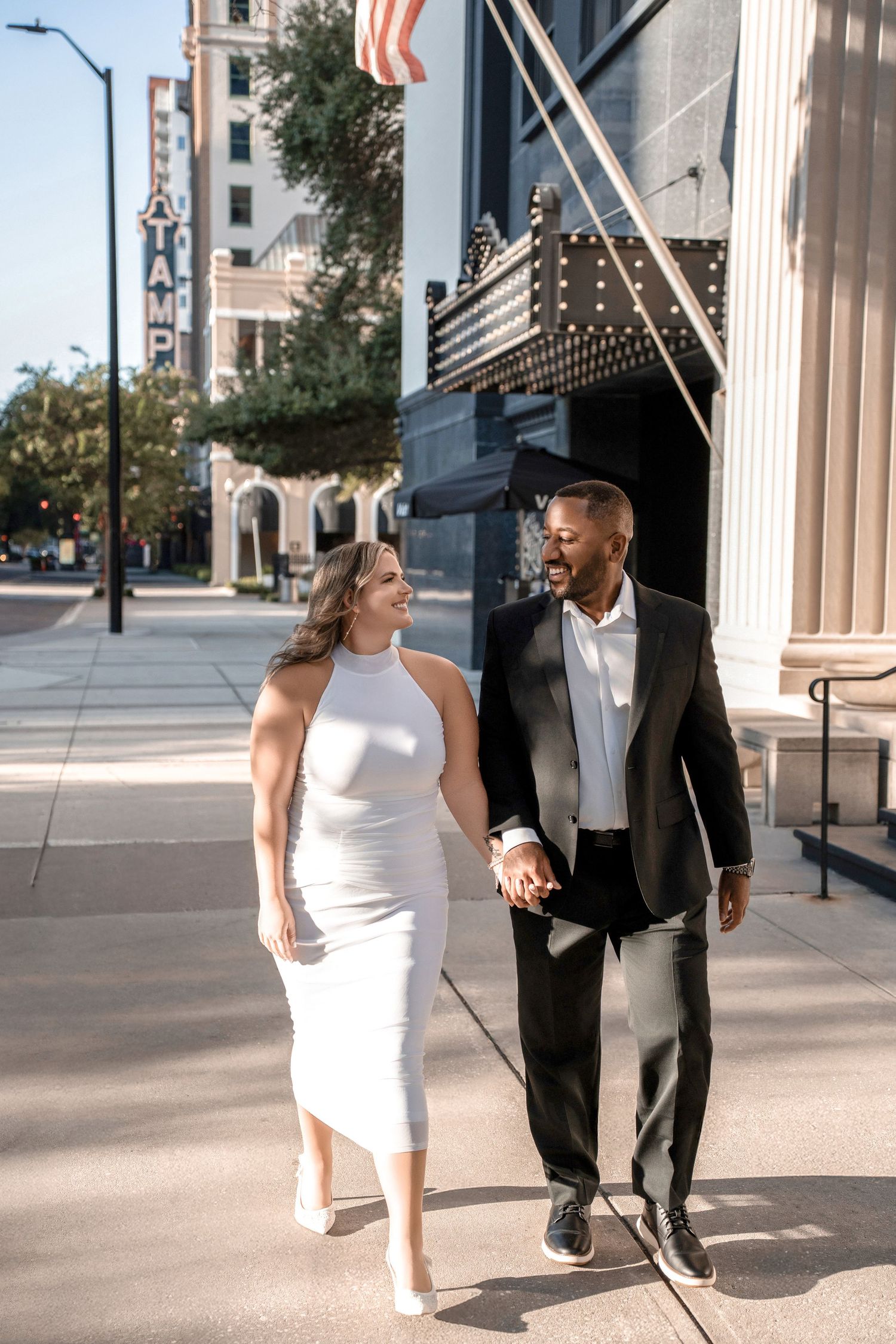 A couple in formal attire walks and poses together between classical white columns in Downtown Tampa