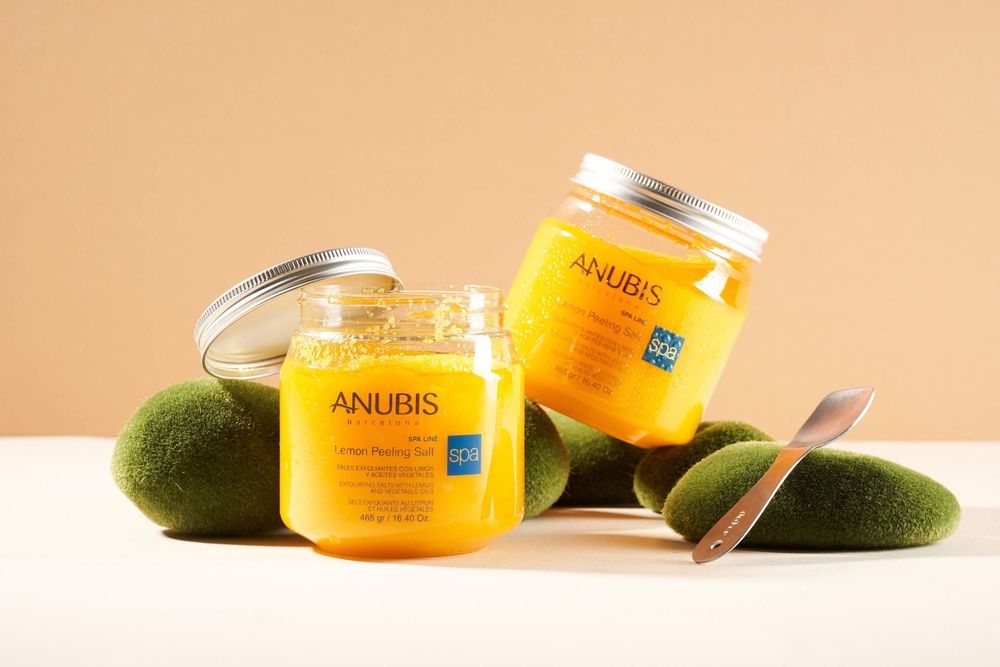 Two yellow Anubis beauty product jars displayed with fresh green avocados and a silver spoon against a beige background.