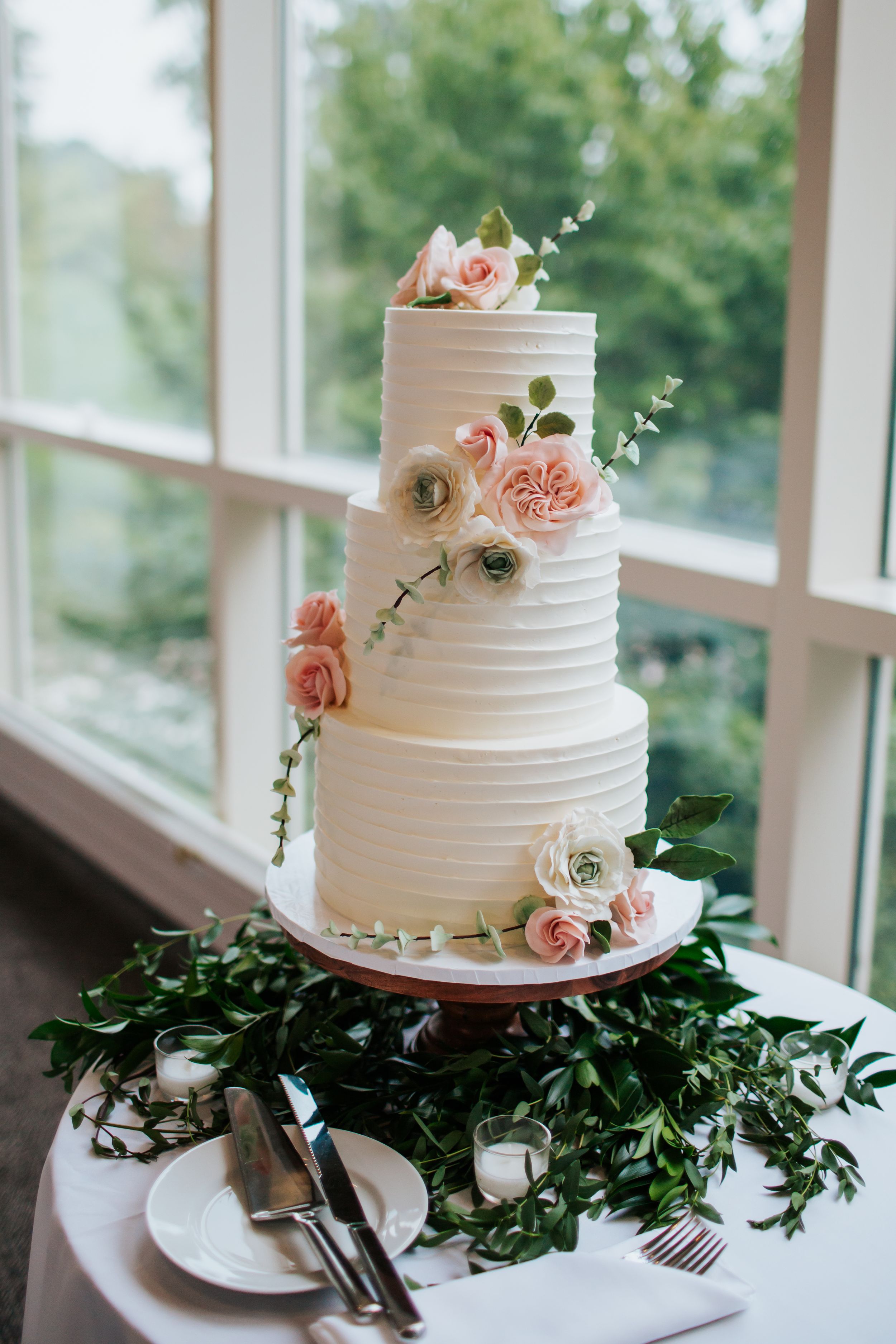 White tiered wedding cake decorated with delicate pink roses sits atop a table adorned with greenery garland.