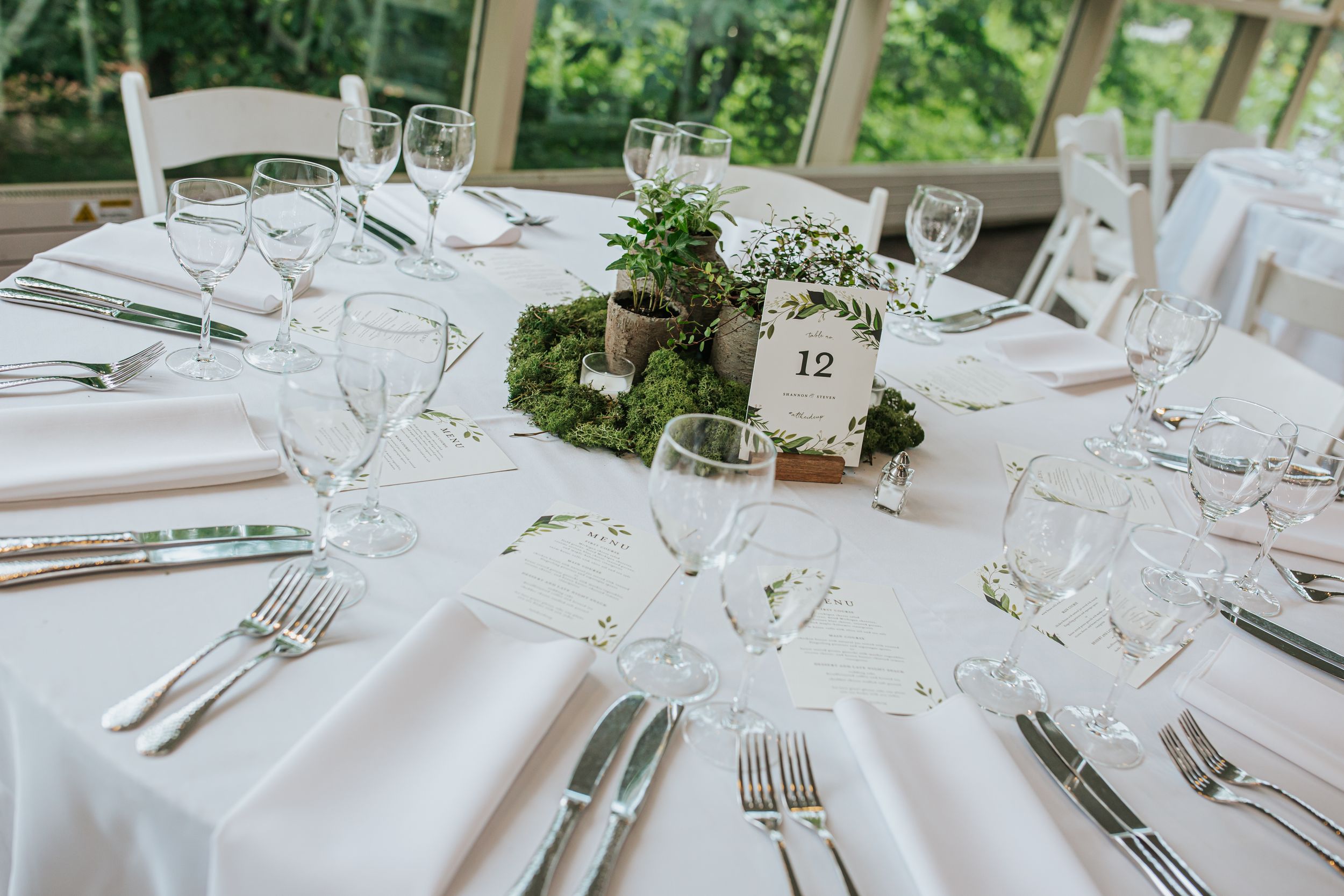 Elegant round wedding table setting with white linens, crystal glasses, and a natural moss centerpiece with greenery.