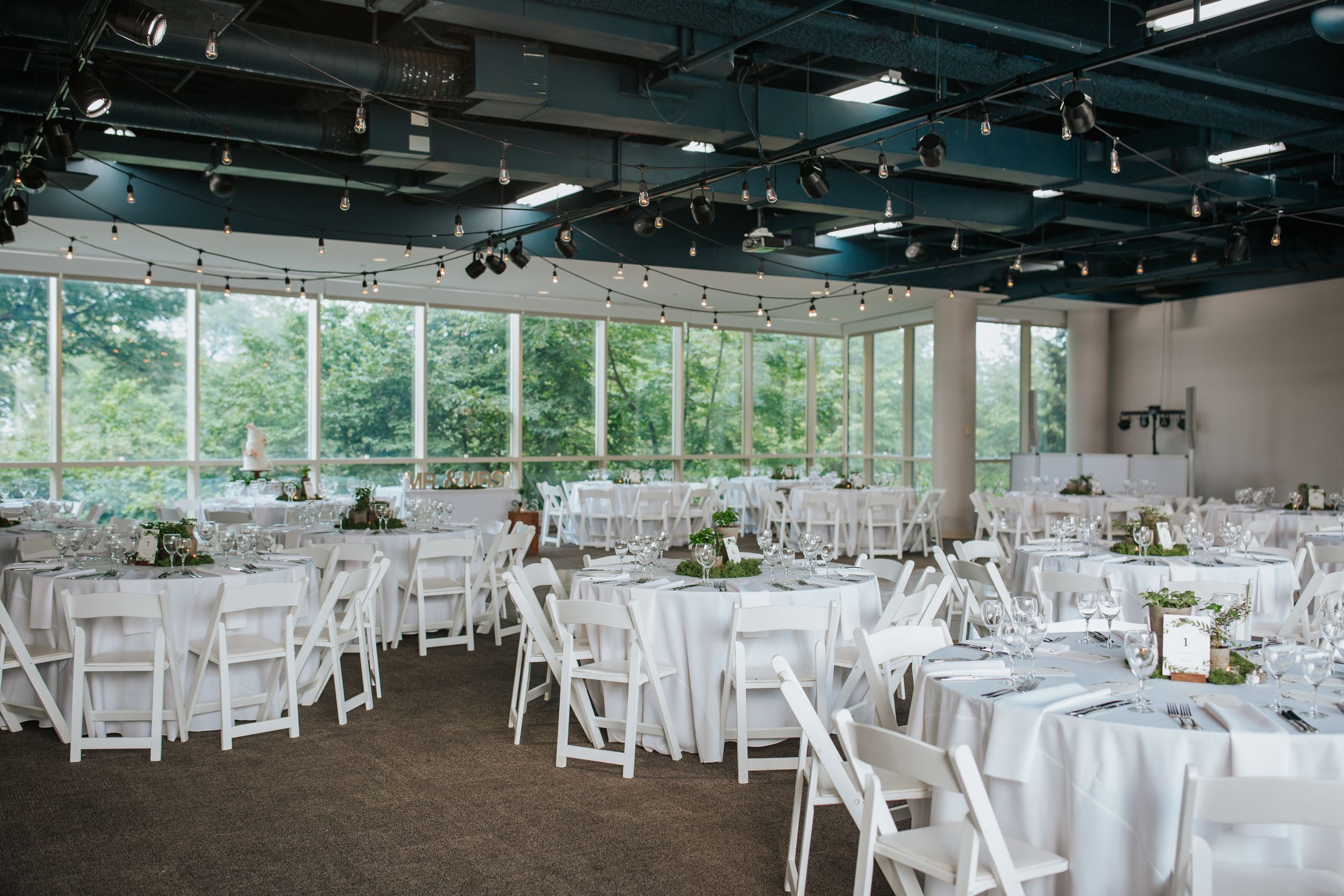 Wedding reception venue setup with white folding chairs and round tables draped in white linens under industrial ceiling.