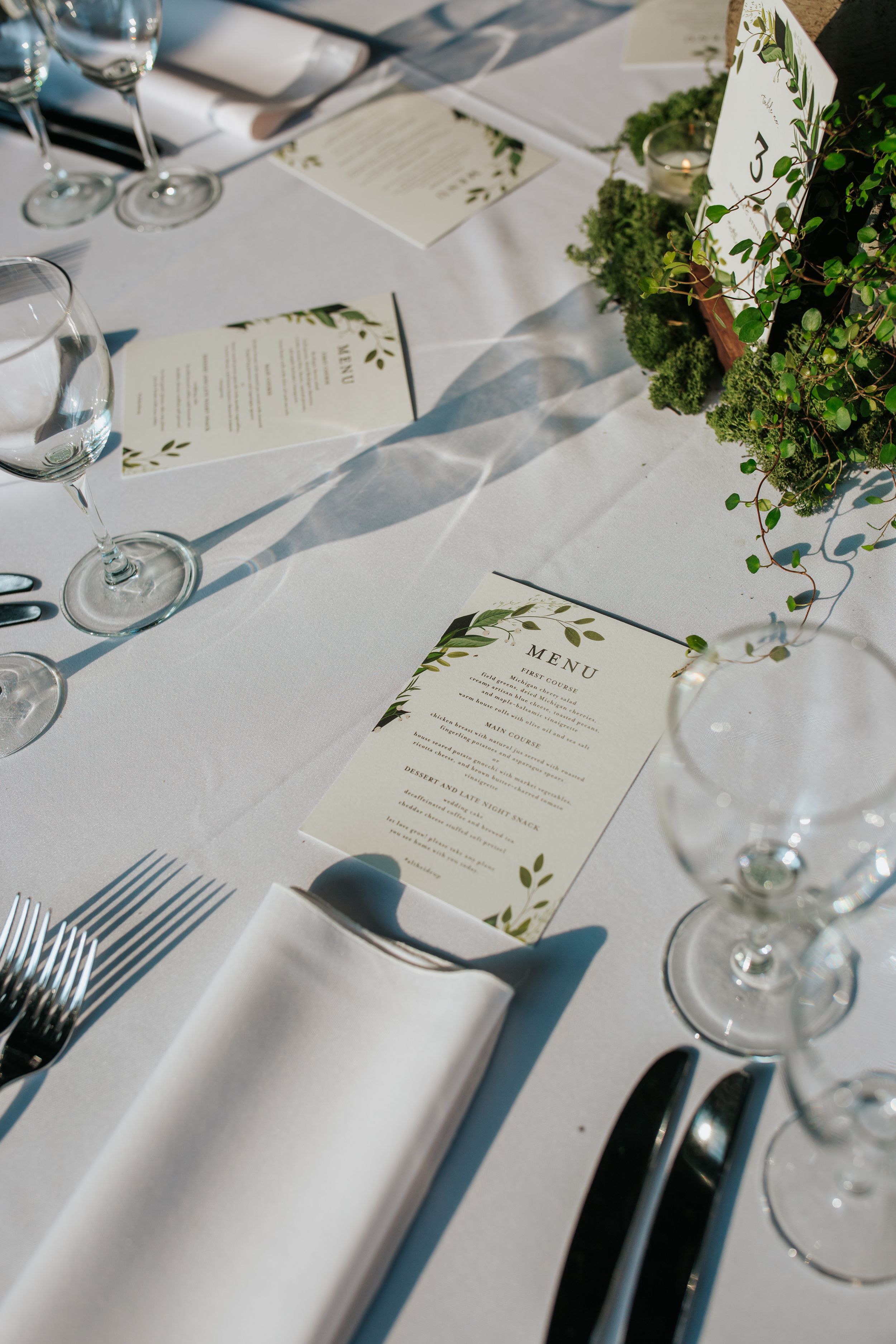 Elegant table settings with white linens, wine glasses, and greenery-decorated menus create a sophisticated dining atmosphere.