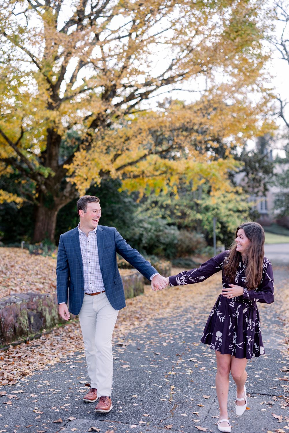 Couple holds hands while walking along leaf-covered path under golden autumn trees.