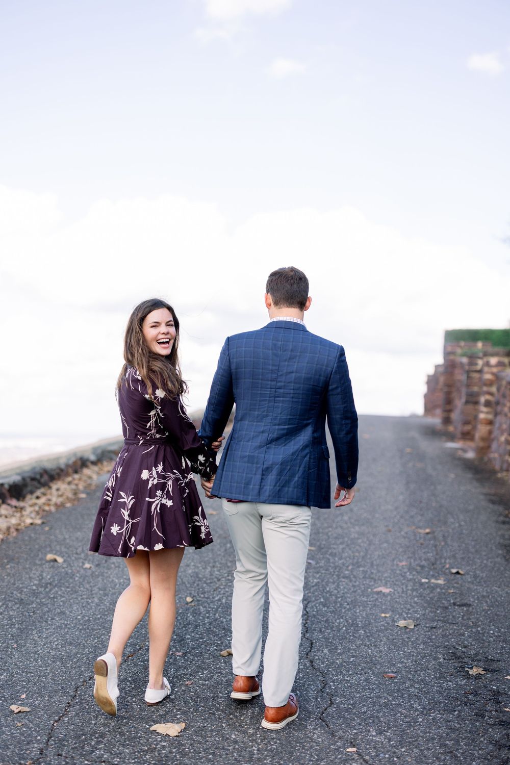 Couple walking on path with stone wall, woman laughing while following behind.