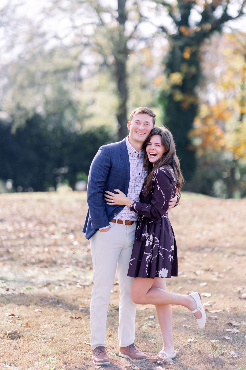 A couple embraces outdoors in autumn, with her wearing a floral dress and him in a navy blazer and khakis.