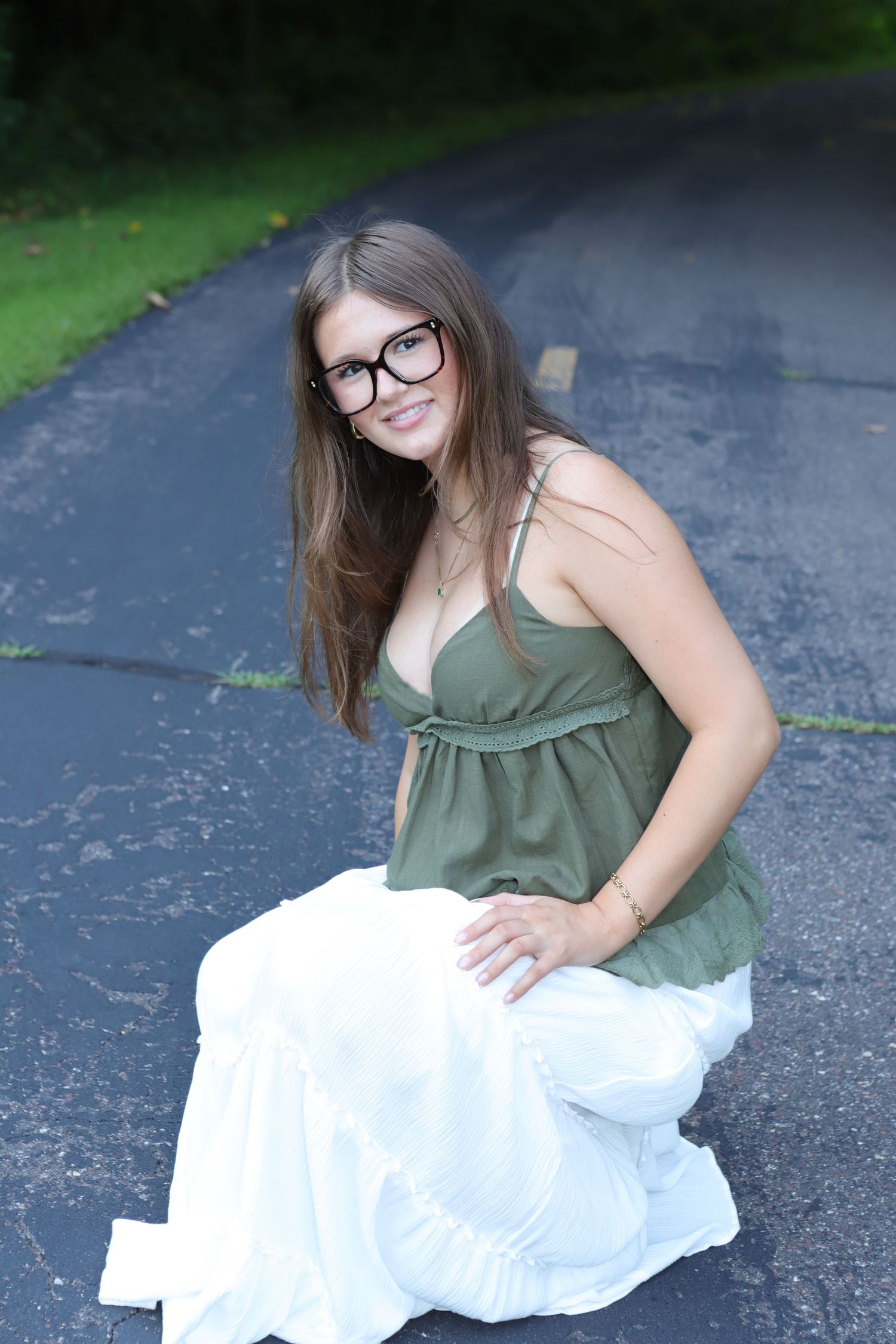Young woman with glasses in green top and white skirt crouching on a driveway, smiling outdoors.