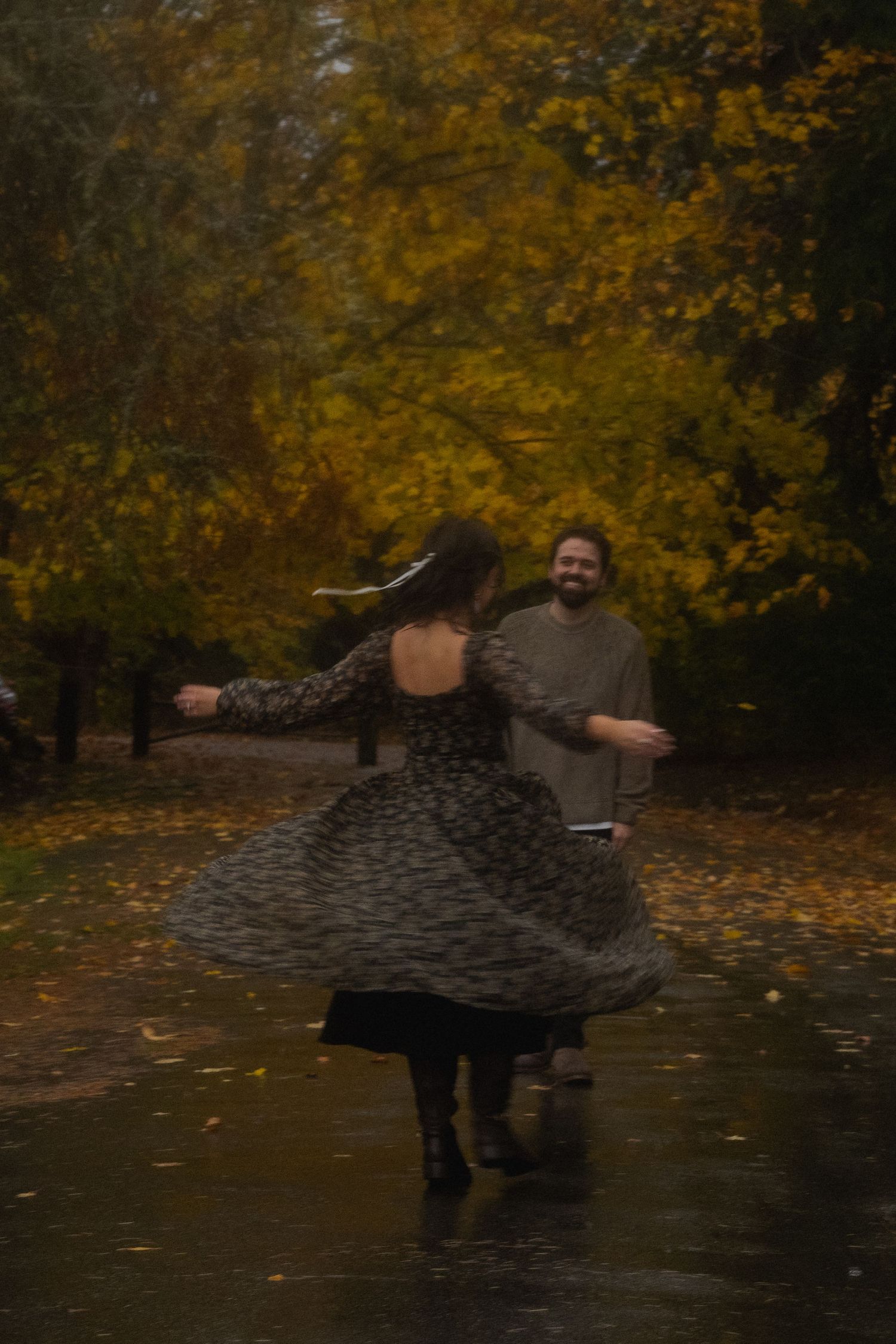 Person in black dress twirls gracefully among autumn trees with golden leaves falling in a park setting.