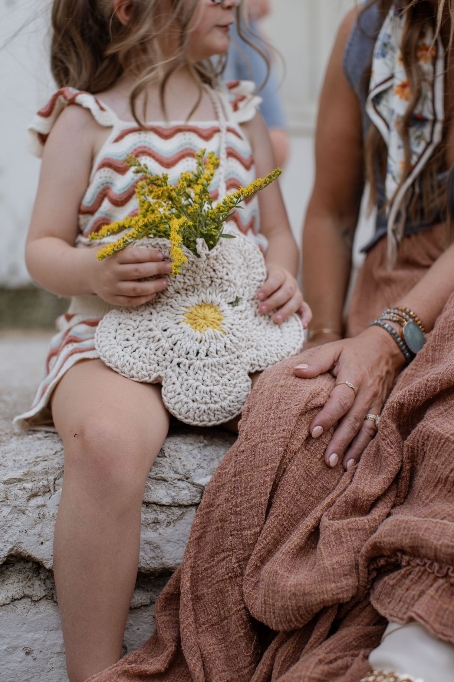 Close-up detail shot of a little girl holding a crocheted flower bag filled with bright yellow wildflowers as she chats with her mom.