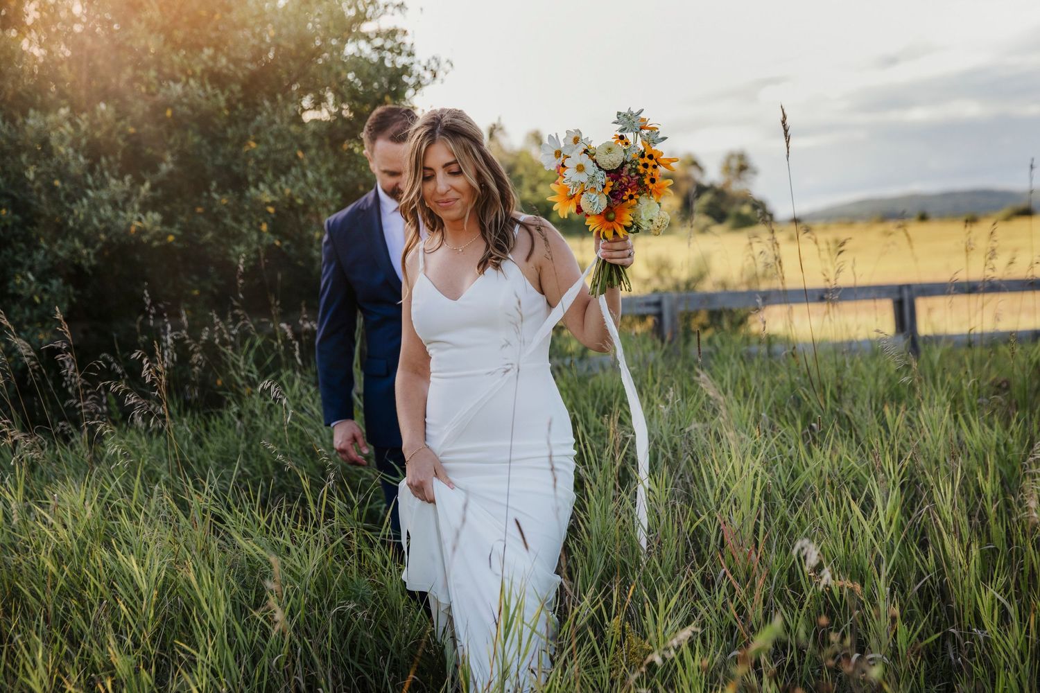 A bride and groom walks together through a golden field of tall grass at sunset during their wedding celebration.
