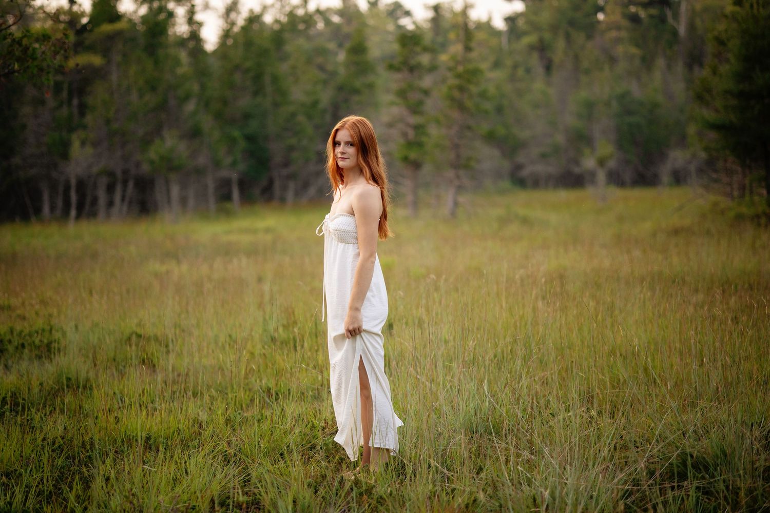 A high school senior girl in a flowing white dress stands in a golden grassy field at sunset with trees in the background.