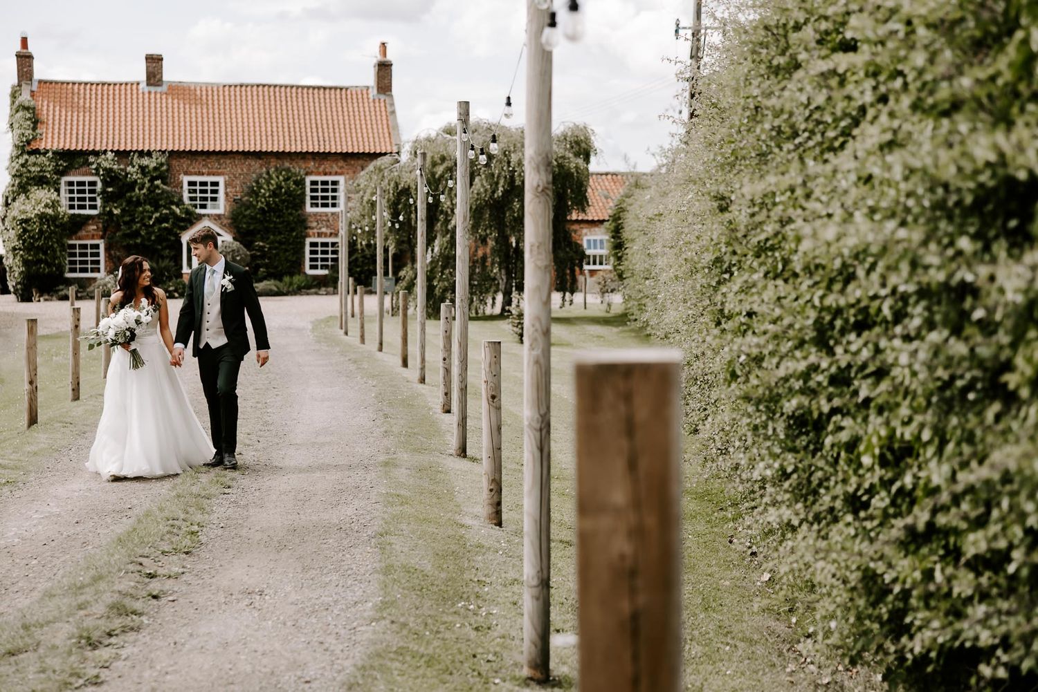 A couple walks down a tree-lined path at a wedding venue with rustic brick buildings in the background.