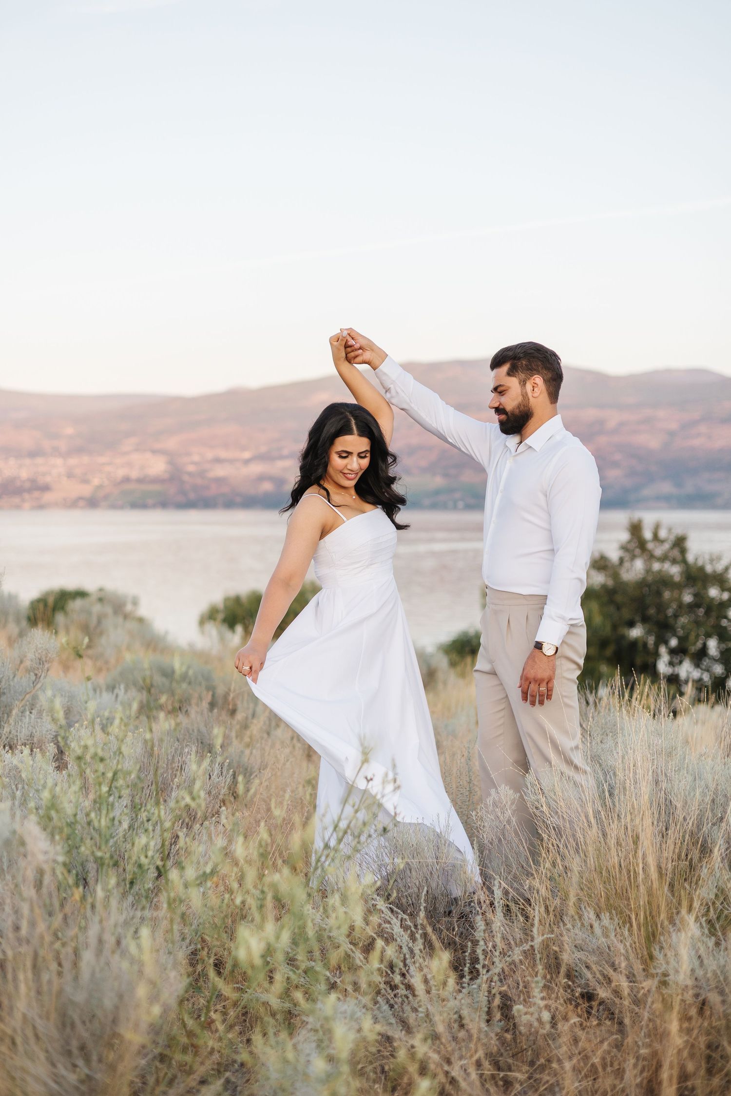 A couple dances together on a hillside overlooking a lake at sunset, with mountains in the background.