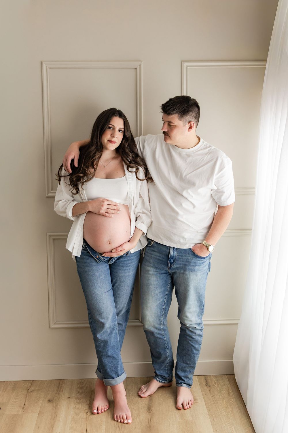 Maternity photo series showing couple in matching white shirts and jeans against white wall.